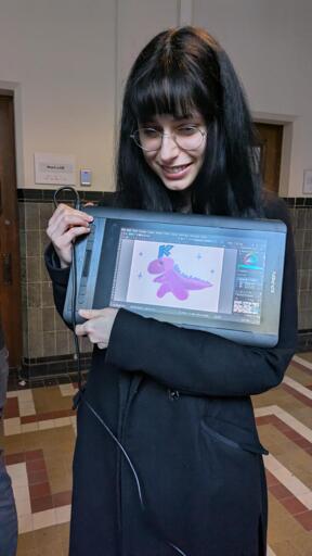 An artist showing off her picture of a pink KDE dragon on a drawing tablet.