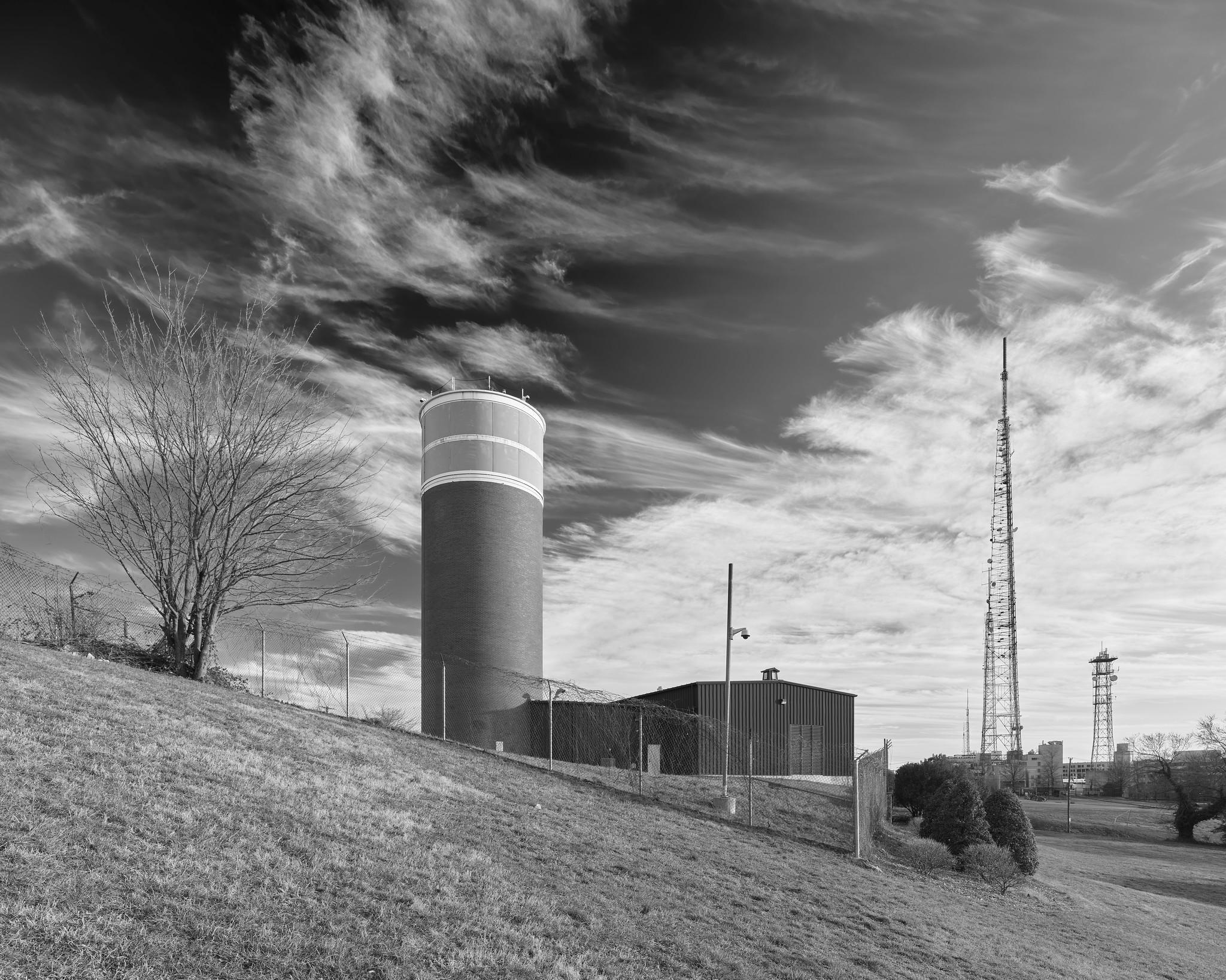 A cylindrical tower, dark brick with a lighter upper section, resembling a water tower or agricultural silo, on a hilltop. Radio towers are visible in the distance. A barren tree is at left.