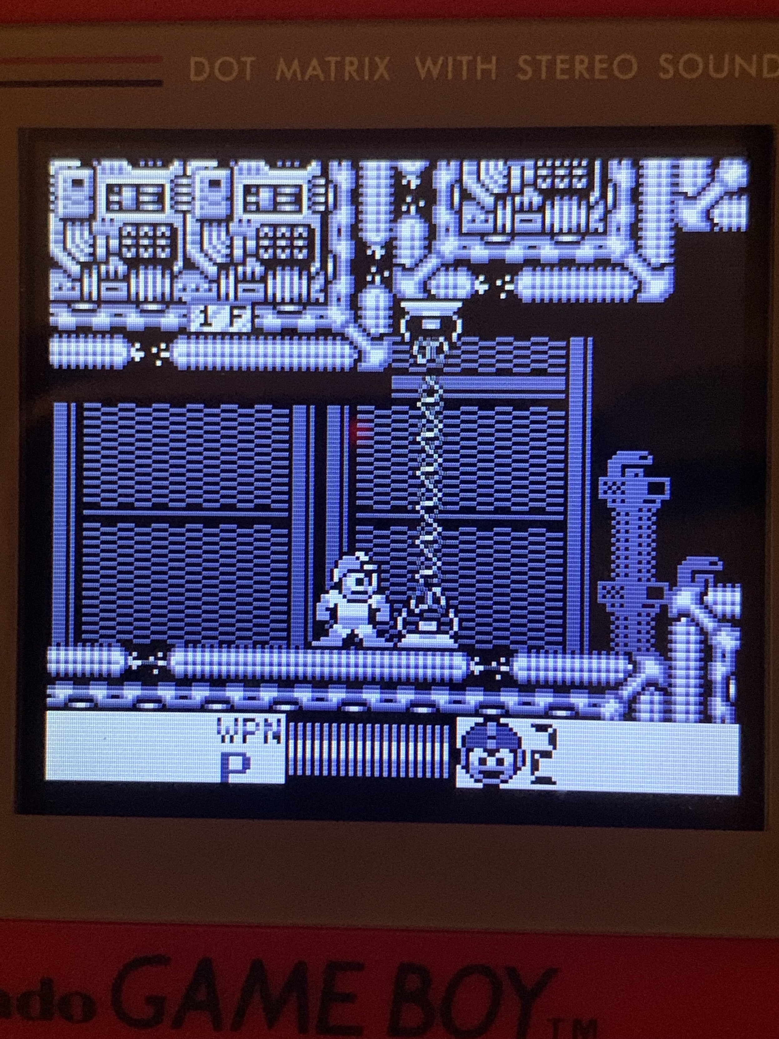 A grayscale display of a retro video game on a Game Boy. The screen shows a character standing near a chain, with various machinery in the background. The status bar indicates weapons and player information at the bottom.