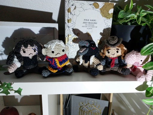 The image shows 5 different small crochet figures. From left to right, the first one is a grumpy looking black haired women, second is a pale person with short white hair and a colorful scarf, third is a person with a sleeveless shirt and a crow's head, fourth is a kind looking man with brown hair, red tie and a cowboy hat, fifth is a blobfish person with a tie and top hat. The figures are all in a sitting position and about 10cm tall.