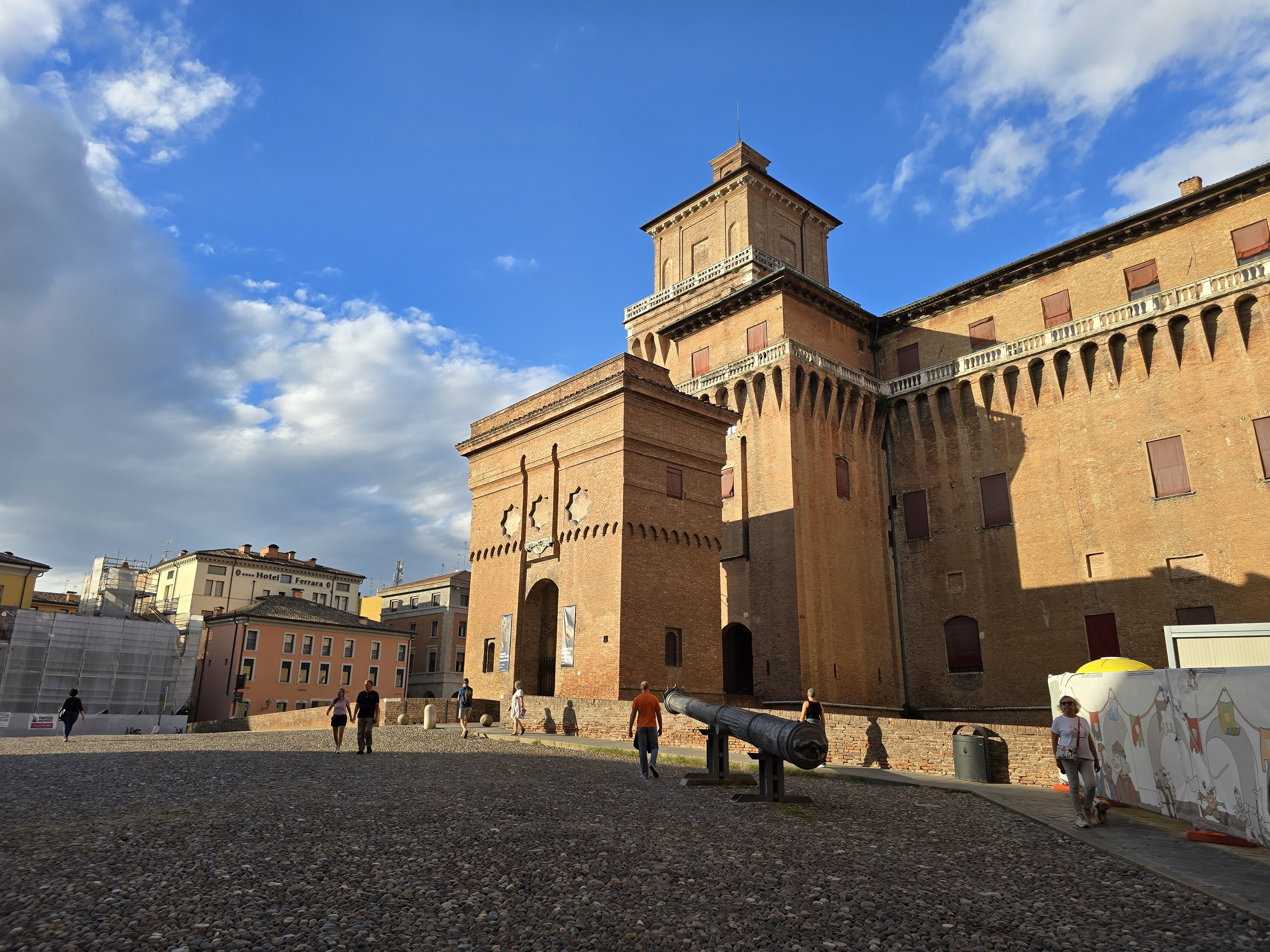 This photo captures the majestic Castello Estense, in Ferrara, Italy, bathed in warm sunlight under a dynamic sky with clouds and patches of clear blue. The imposing medieval structure, built from red brick, showcases its robust towers, arched windows, and crenellated walls. The cannon displayed in the foreground adds a historical touch, while a few people stroll leisurely across the cobblestone square, contributing to the serene yet grand ambiance. In the background, modern buildings, including a hotel, subtly blend into the scene, emphasizing the juxtaposition of history and modernity.
