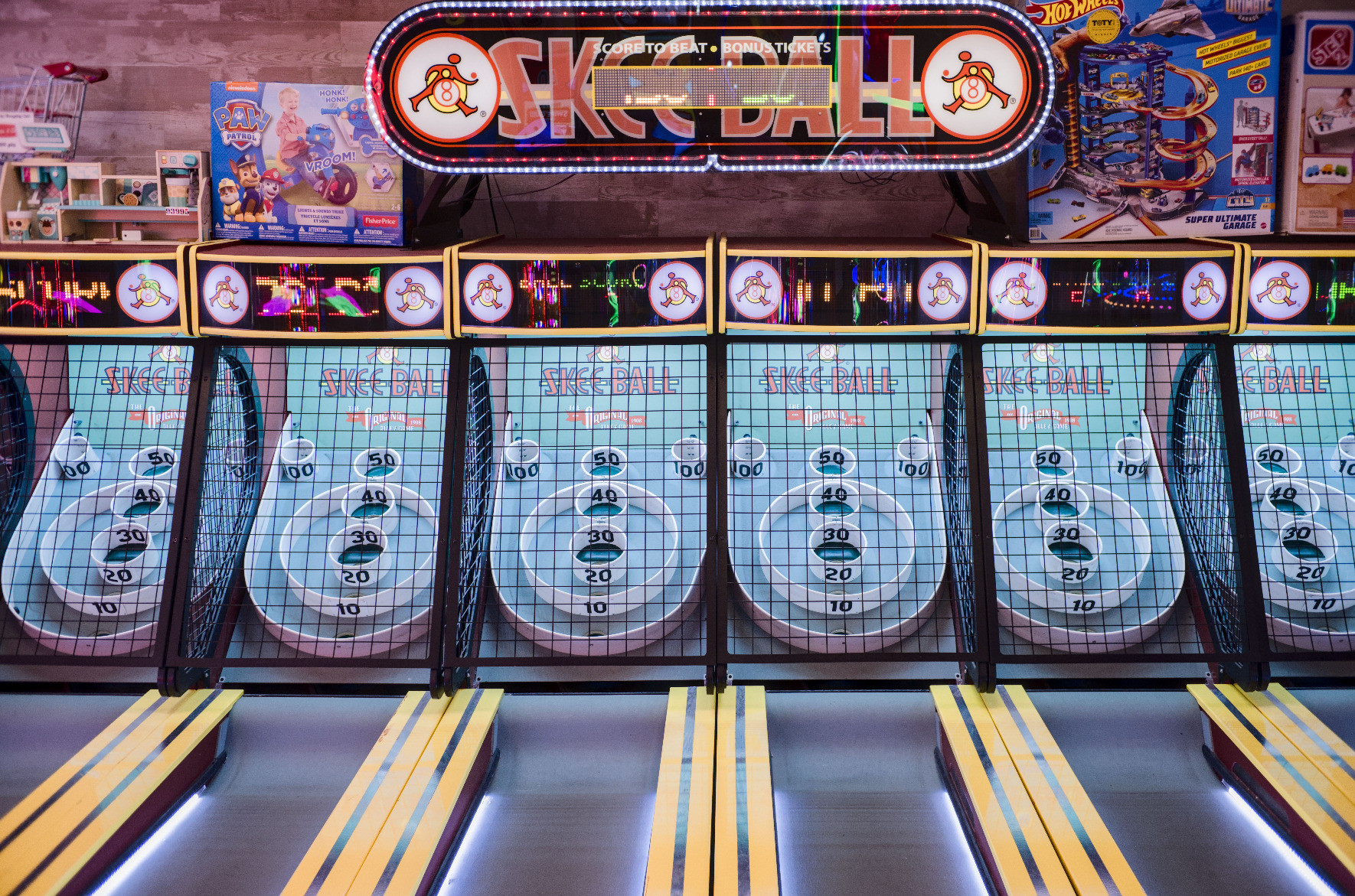 A photo of 6 skeeball lanes with a big SKEEBALL sign overhead. The lanes are neon yellow and the skeeball targets are light blue. The photo has major retro-arcade vibes.