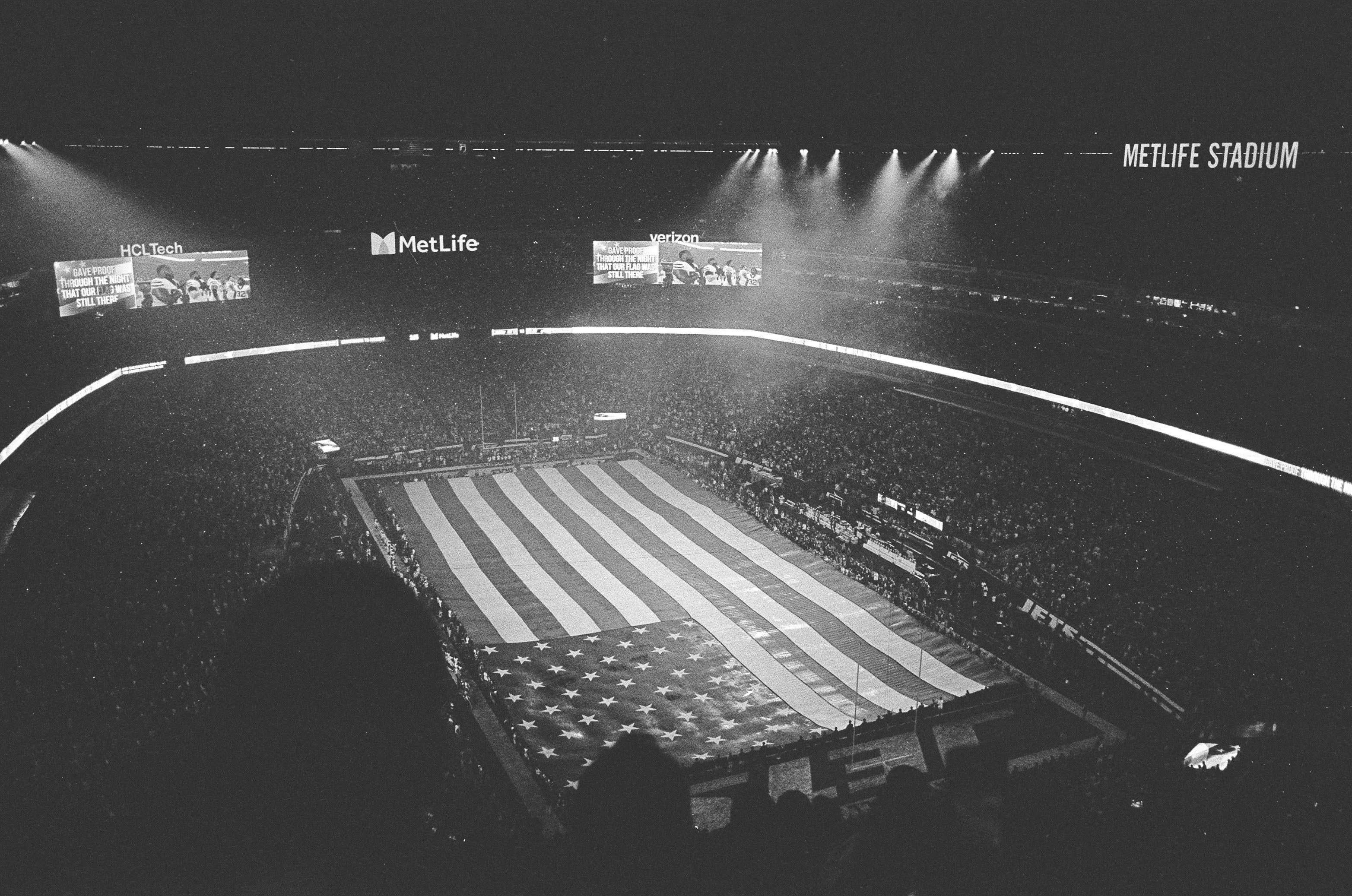 A black and white photo from the top of met life stadium. An American flag covering the entire field is shown. Primarily lit by the directional floodlights at the top of the stadium. 

Taken on a Nikon N70 with a 28mm lens. Film is Ilford HP5. 