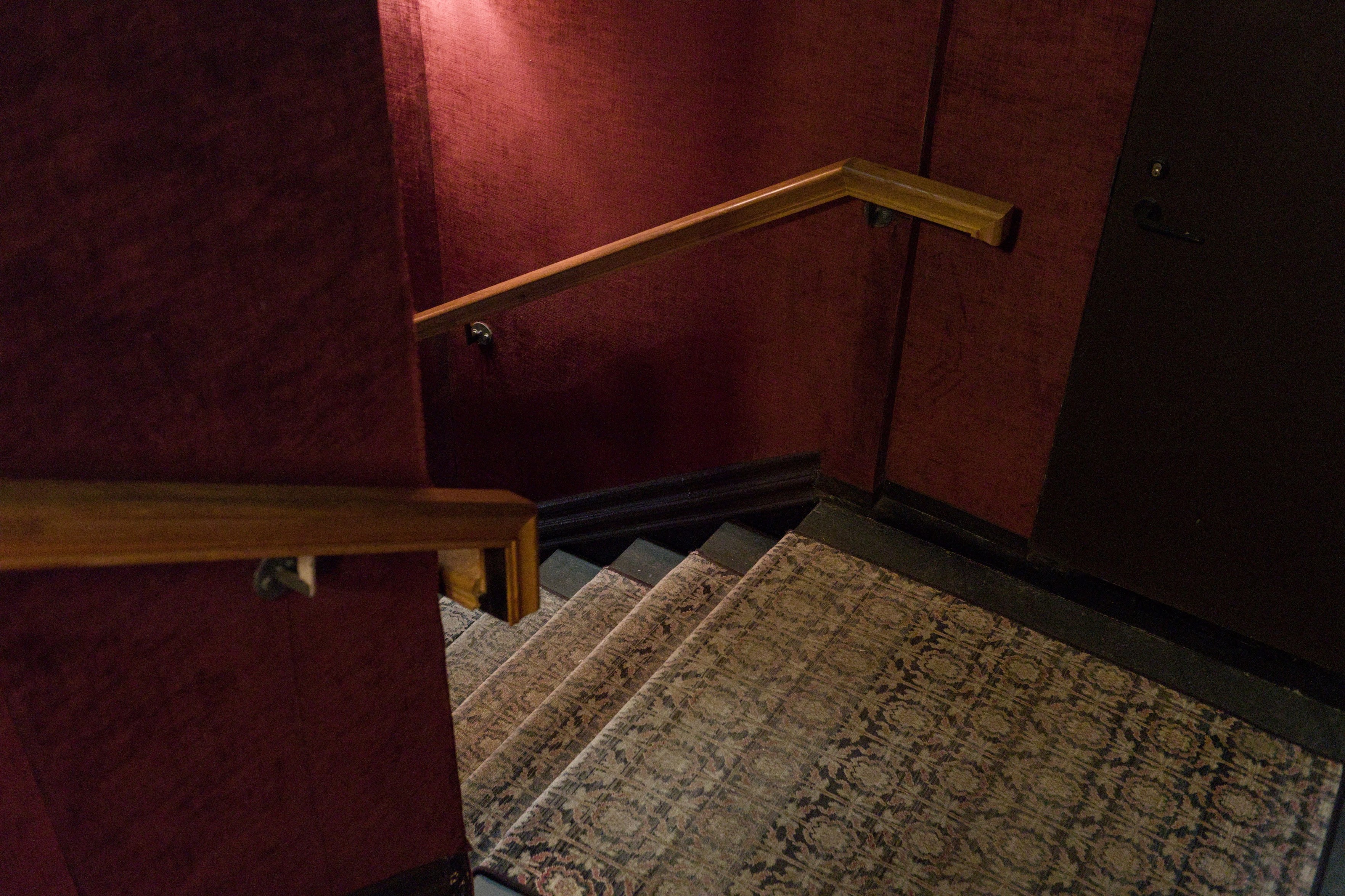 A color photo of the stairwell at Fotografiska. A sconce lights the red walls and old carpet dimly. 