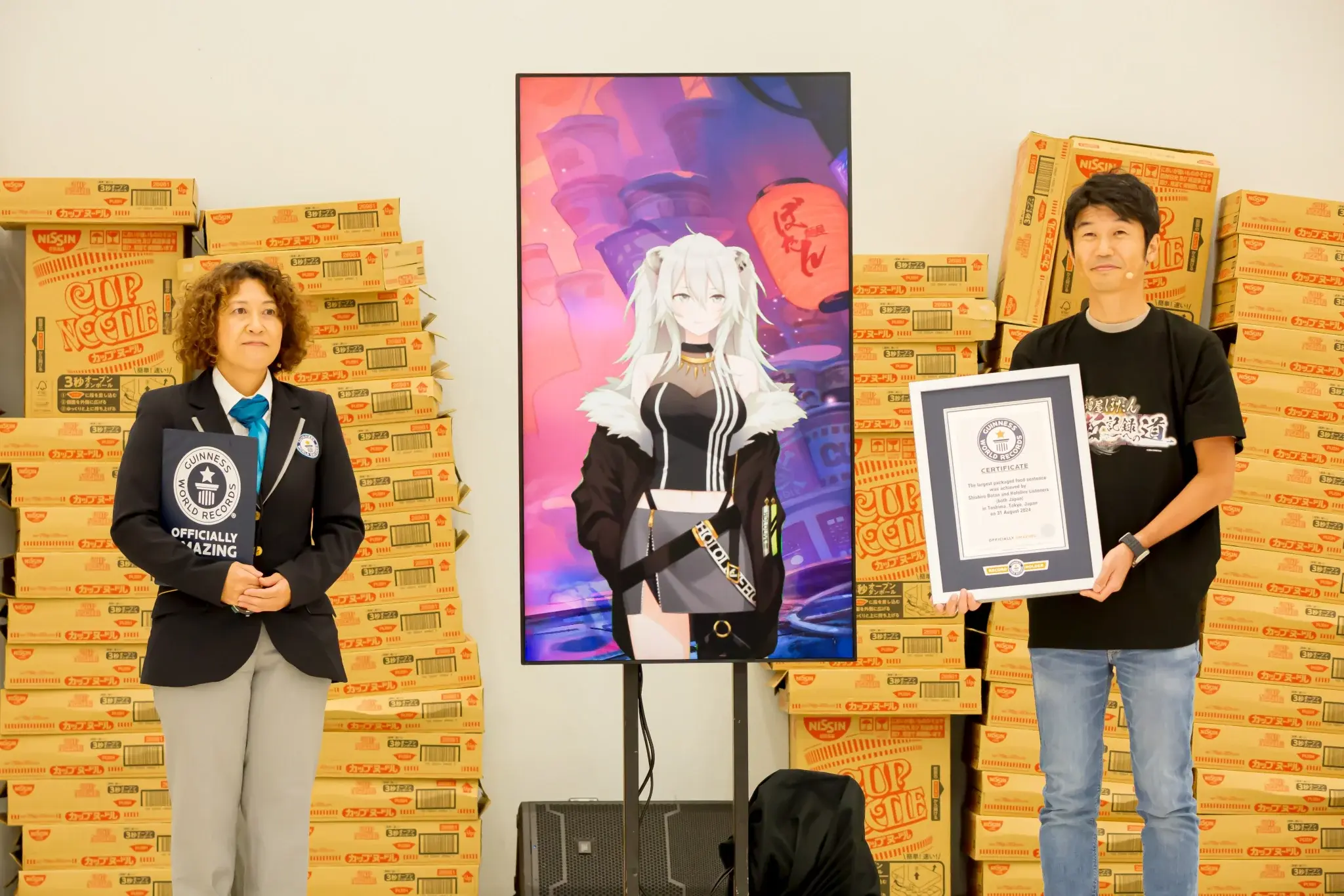 Shishiro Botan on a large vertical screen, flanked by a Guiness World Records official and Yagoo, who is holding a framed Guiness World Records certificate. Behind them are large stacks of boxes for cup noodles.