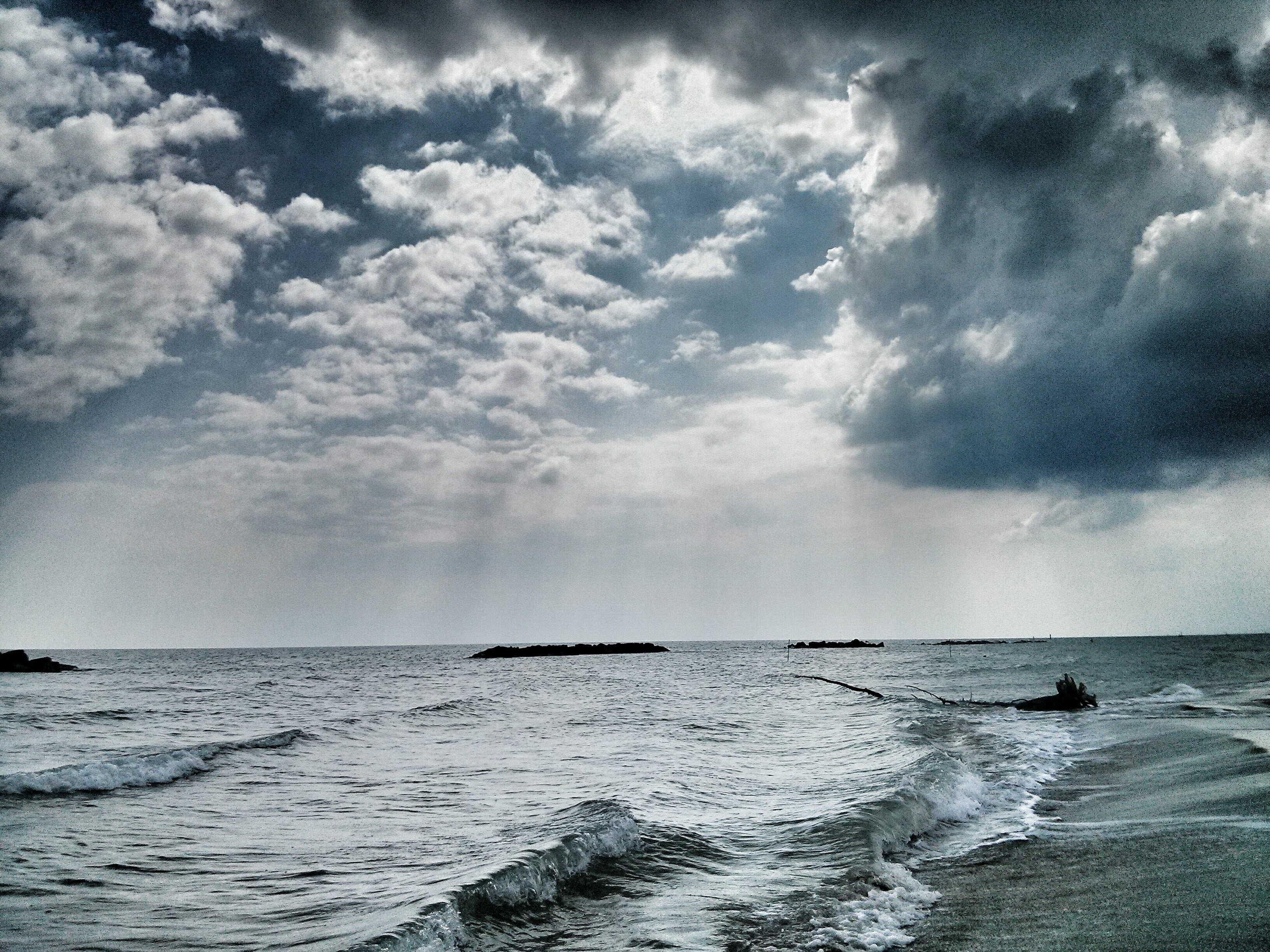 A peaceful seaside view under a dramatic, overcast sky, with a mix of heavy clouds and sunlight streaming through gaps. The waves gently lap at the shore, creating a soft, rhythmic sound that contrasts with the looming clouds. Despite the strength of nature's presence, the scene evokes a perfect sense of silent solitude, as if the power of the elements enhances the serenity of this quiet Sunday moment. Driftwood floats in the distance, further emphasizing the natural flow of time and calmness.