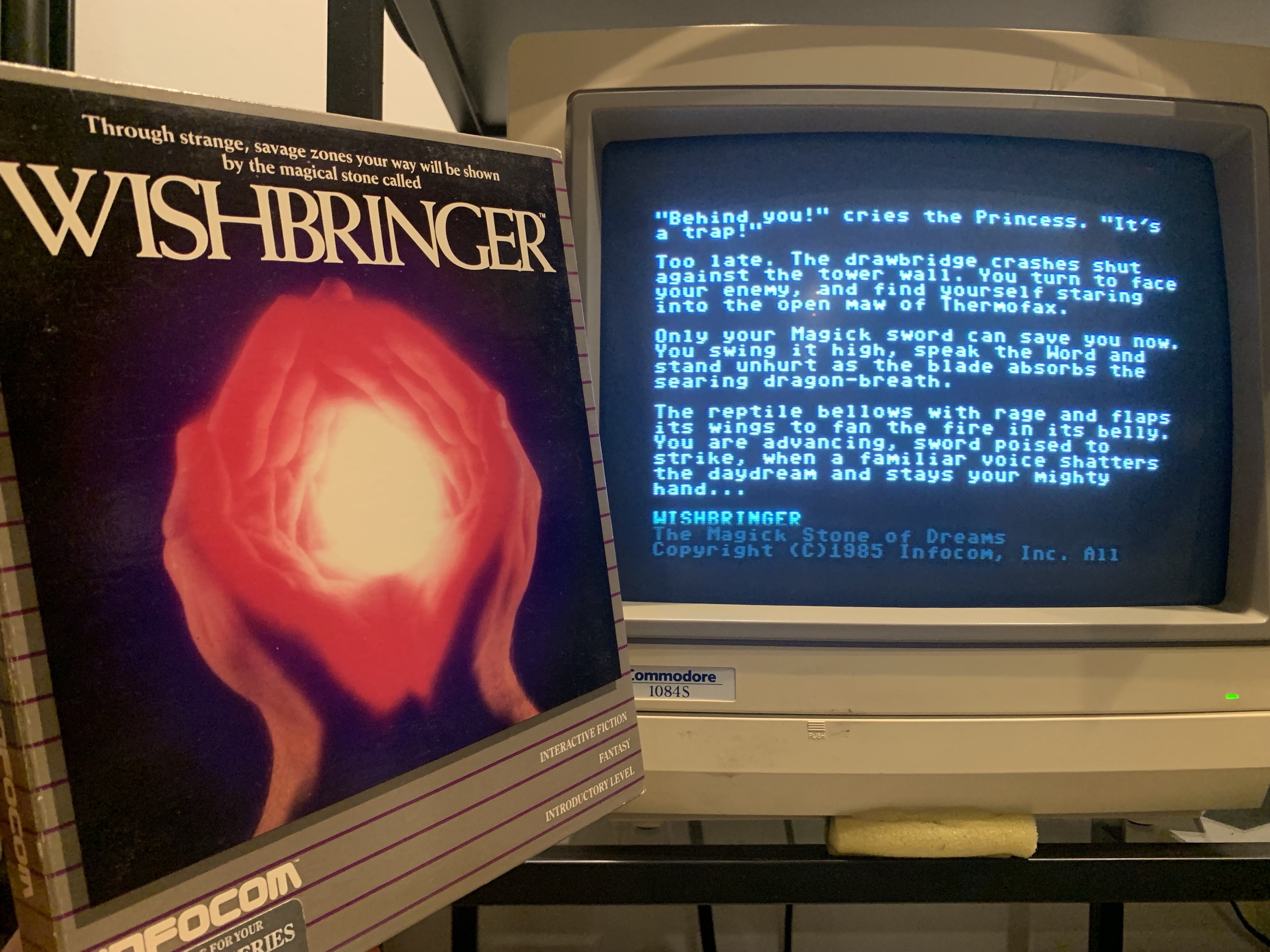 The image features the box art for the interactive fiction game "Wishbringer" by Infocom, alongside a vintage computer monitor displaying text from the game. The box art depicts two hands surrounding a glowing orb, while the monitor shows a narrative from the game