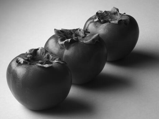 Three persimmons, arranged diagonally in the frame, on a blank white surface.