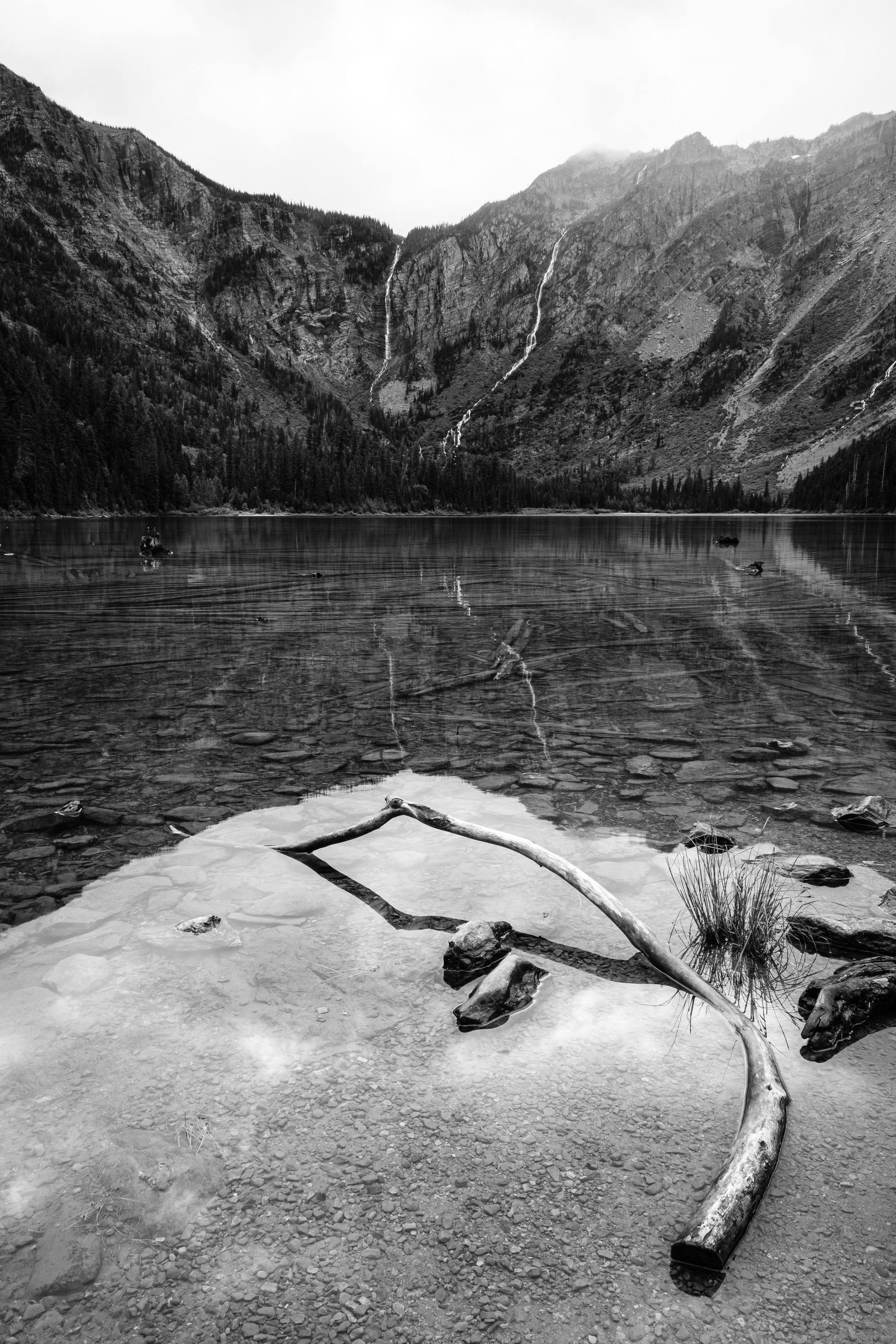 Avalanche Lake. In the foreground, a few rocks and a curved log half submerged in the lake's water. In the background, two waterfalls running down the sides of the mountain at the end of the lake.