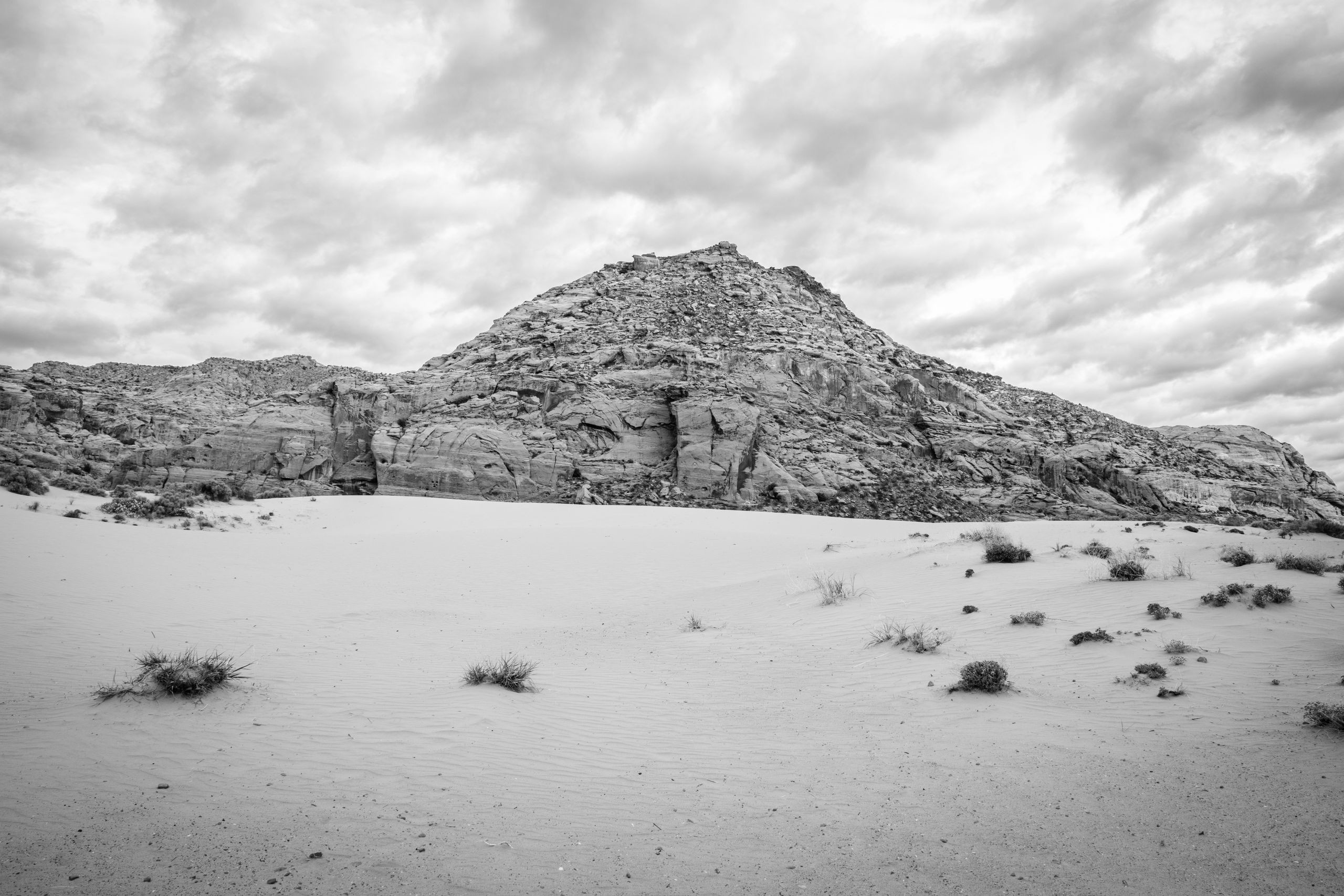 A rocky cliff in Snow Canyon State Park, pictured under cloudy skies behind sand dunes with vegetation.