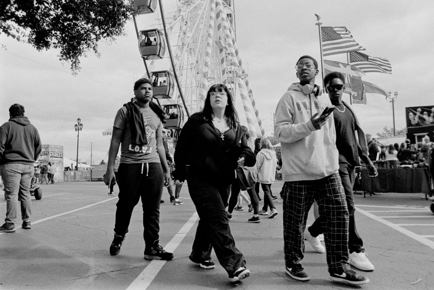 A black and white photo of a group of young people at the NC state fair. The photo is taken from a low perspective and a large ferris wheel and flags blowing in the wind are in the background.

Nikon N70 28mm 
Cinestill BW XX