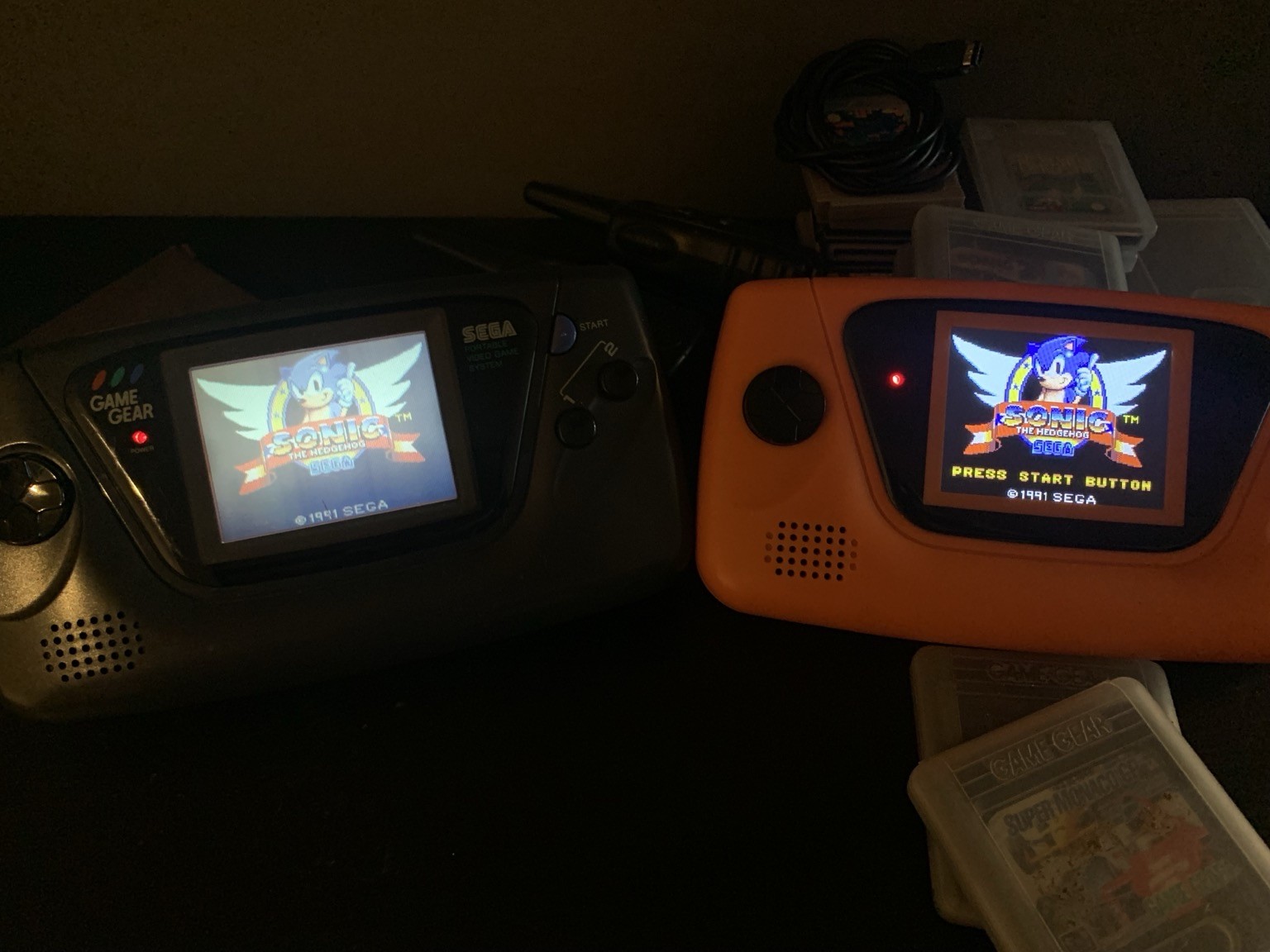 Two GameGear handheld consoles. The left one is black and has a washed out LCD screen showing the Sonic 1 title screen. The right one is orange with black buttons and shows the same screen with crisp colors and excellent contrast. 