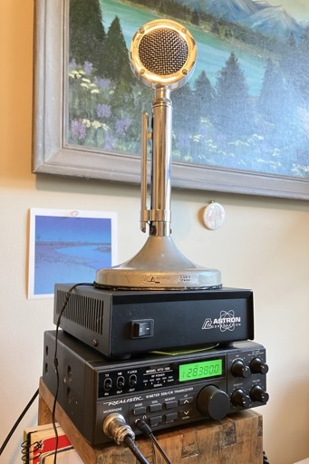 A small rig of ham components stacked on a wooden shelf. Top to bottom: old timey looking chrome microphone, black power supply, and gray mobile transceiver