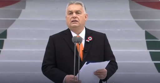 Il primo ministro ungherese Viktor Orbán, davanti a un microfono, tiene in mano in mano un foglio di carta durante una cerimonia ufficiale. Si sta presumibilmente rivolgendo alla folla. Appuntata sul bavero del suo vestito c'è una coccarda con i colori dell'Ungheria. Immancabile la cravatta arancione, colore distintivo di Fidesz, il suo partito. 