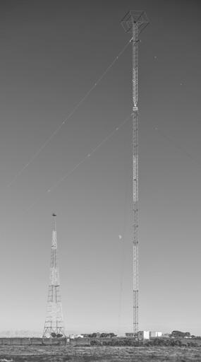 Two radio antenna masts in a field. The taller mast, on the left, is a guyed tower with a hexagonal structure at the top. The shorter mast on the right, toward the background, is freestanding.