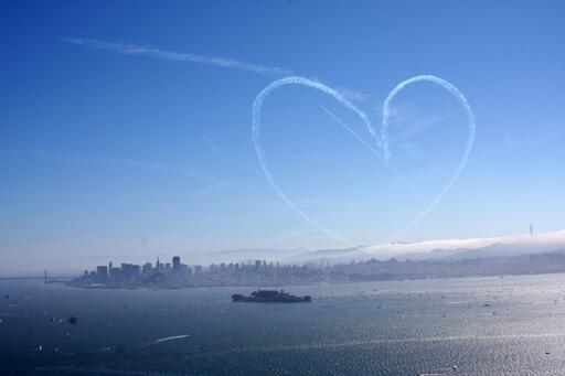 An aircraft's smoke trail has left a heart having in the sky over San Francisco, and the plane adds a fresh arrow through it. The city has partial fog at the view is from the top of Angel Island, with Alcatraz Island in the foreground. Sutro tower peeks up through the fog on the right.