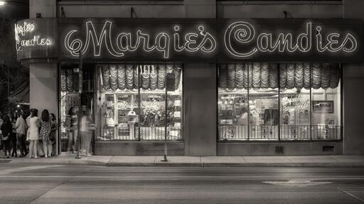 Exterior of an old-school sweets shop at night. A neon sign above the windows reads "Margie's Candies" in a stylized script. A small line of people wait at the door.