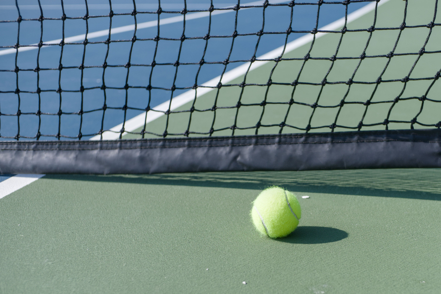 A color photo of a tennis ball laying in front of the net on a green and blue court.