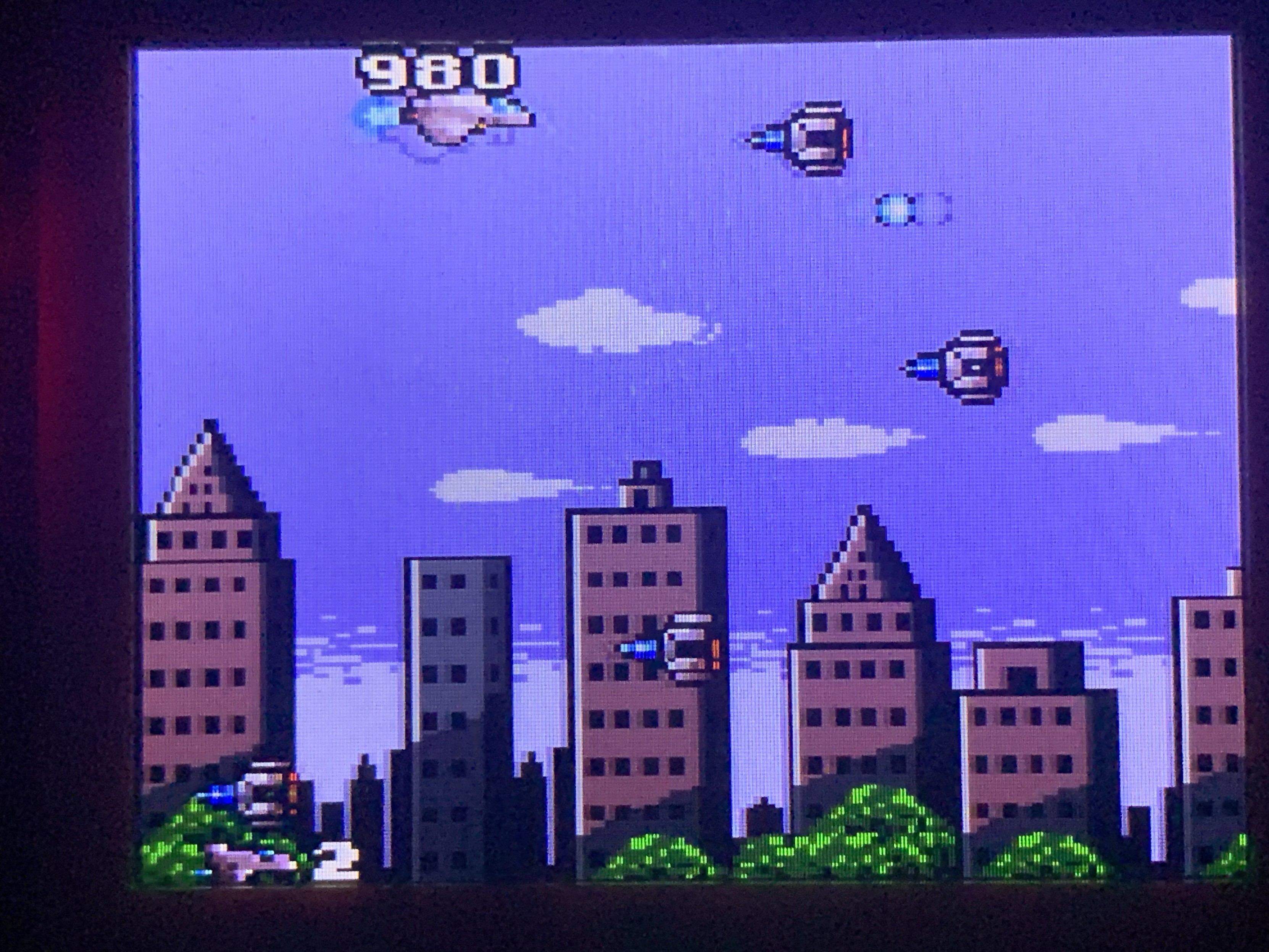 A 2D scene showing highrise buildings in a city. Spaceships coming in from the right while the player‘s plane is at the top left. 